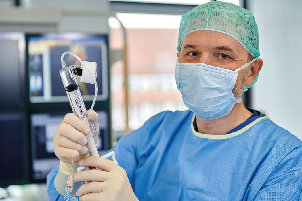 Männliche Person in blauer OP-Arbeitskleidung mit Mundschutz und OP-Haube.