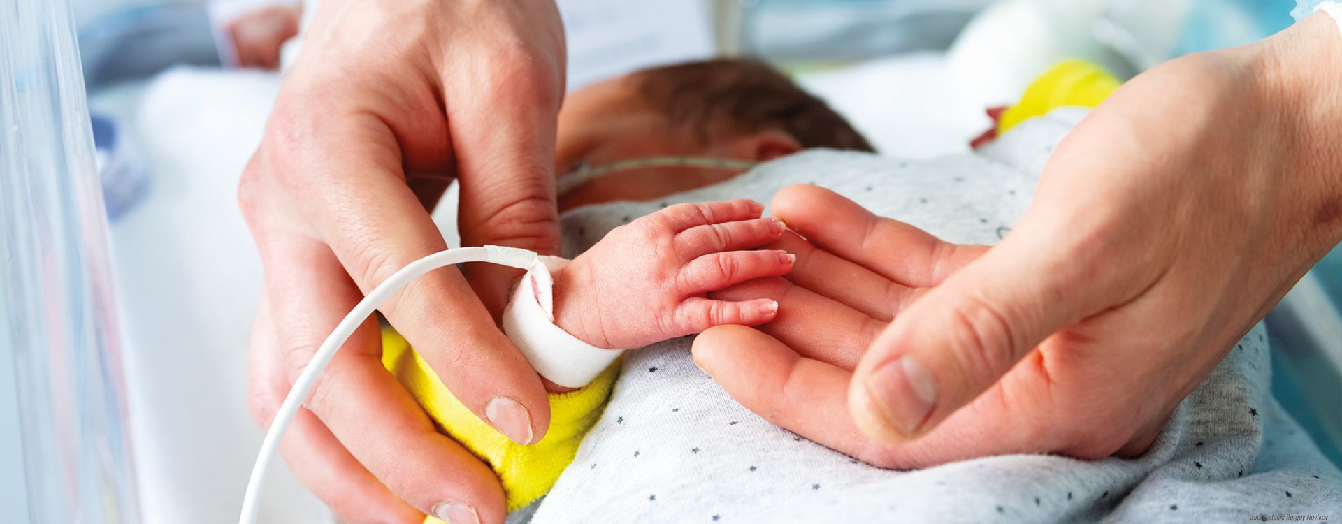 Zwei Hände halten die winzige Hand eines Frühgeborenen im Wärmebett, das Neugeborene ist an verschiedene medizinische Geräte angeschlossen