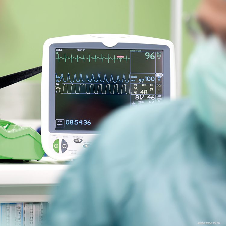 unscharf dargestellte Person in grüner Krankenhausarbeitskleidung vor einem Überwachungsmonitor