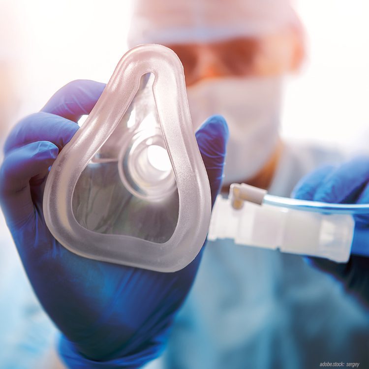 unscharf dargestellte Person in OP-Arbeitskleidung zeigt eine Beatmungsmaske