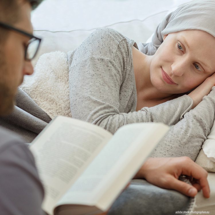 Liegende Krebspatientin mit Kopftuch bekommt von ihrem Partner aus einem Buch vorgelesen.