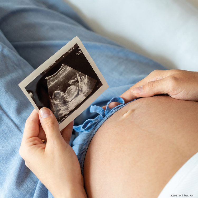 liegende Frau mit Schwangerschaftsbauch betrachtet Ultraschallbild ihres Babys und hält eine Hand auf ihren Bauch