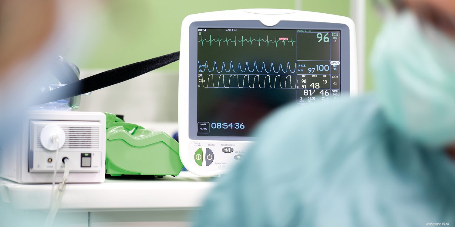 unscharf dargestellte Person in grüner Krankenhausarbeitskleidung vor einem Überwachungsmonitor