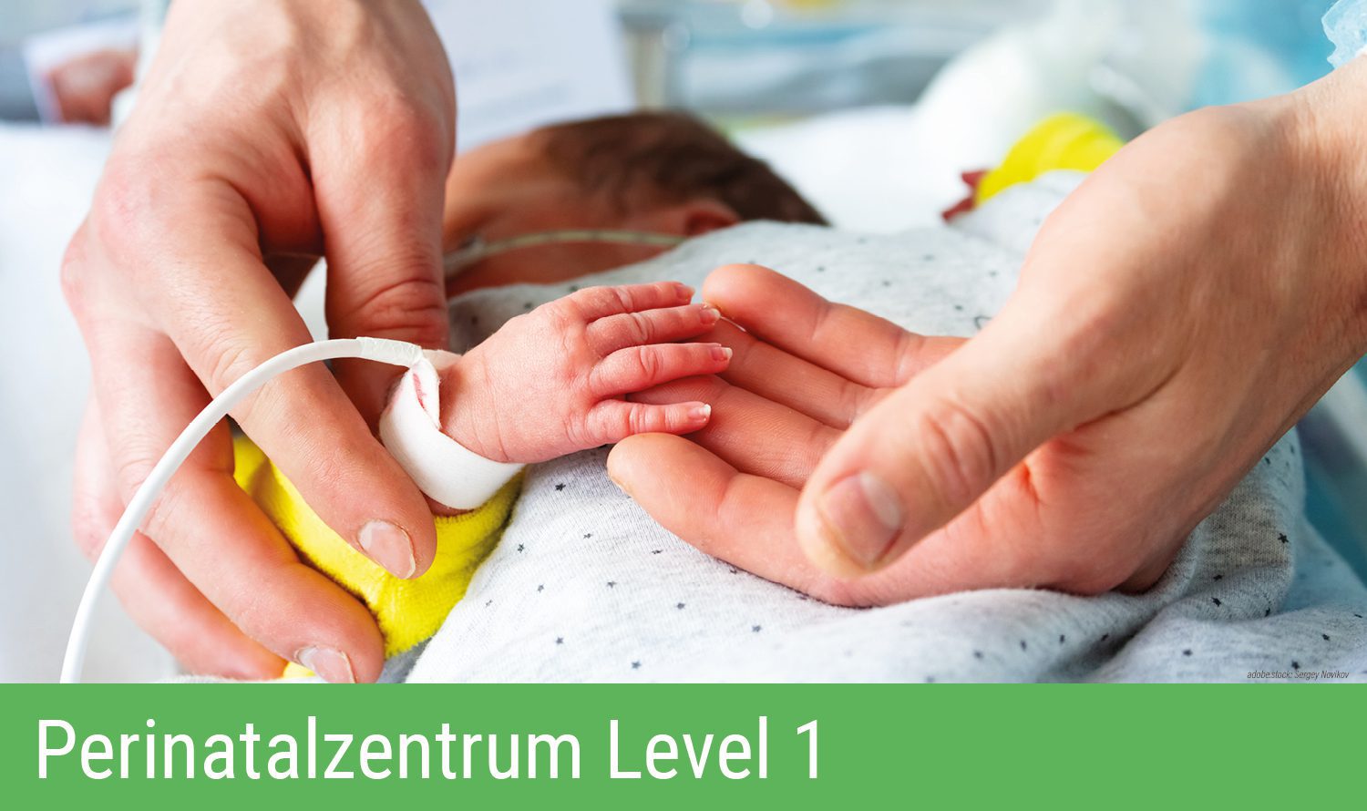 Zwei Hände halten die winzige Hand eines Frühgeborenen im Wärmebett, das Neugeborene ist an verschiedene medizinische Geräte angeschlossen. Grüner Balken am unteren Bildrand mit Aufschrift Perinatalzentrum Level 1.
