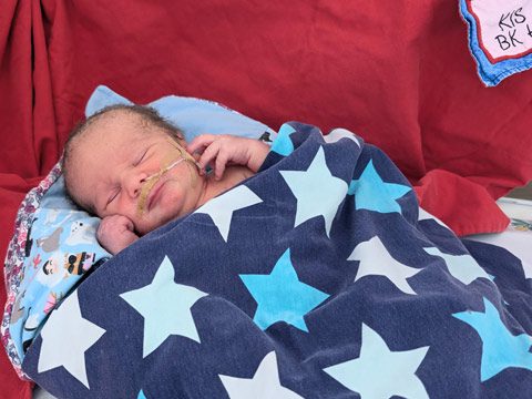 Neugeborenes Timofey mit Nasenkanüle liegt in blauer Sternen-Decke eingewickelt auf rotem Untergrund im Krankenhaus